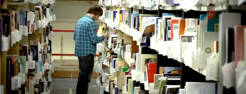 A WeBuyBooks warehouse worker picking stock from tall shelves packed with second-hand books