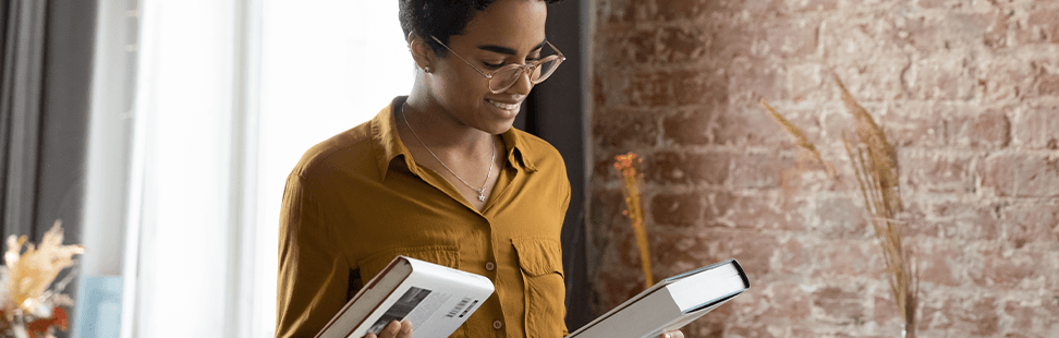 Woman in a mustard shirt smiling as she looks through a stack of hardback books at home