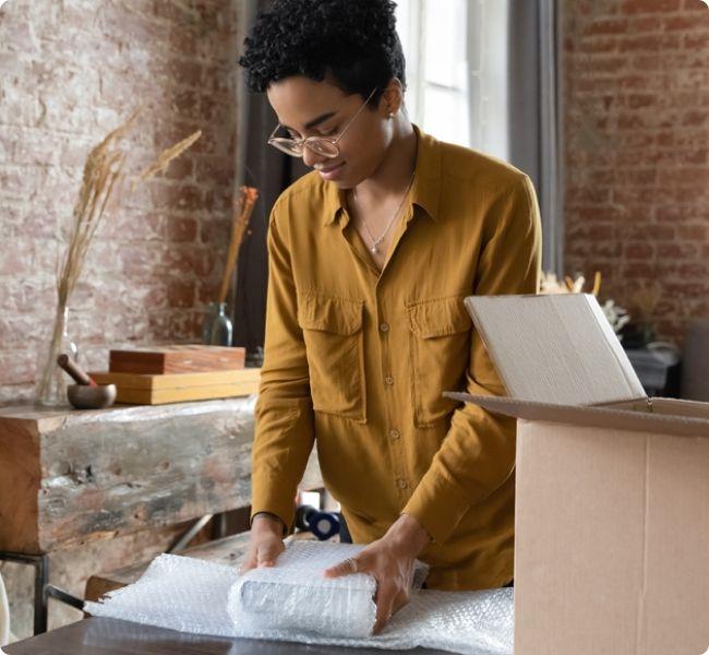 Woman wrapping an item in bubble wrap beside an open cardboard box, preparing to send in her collection