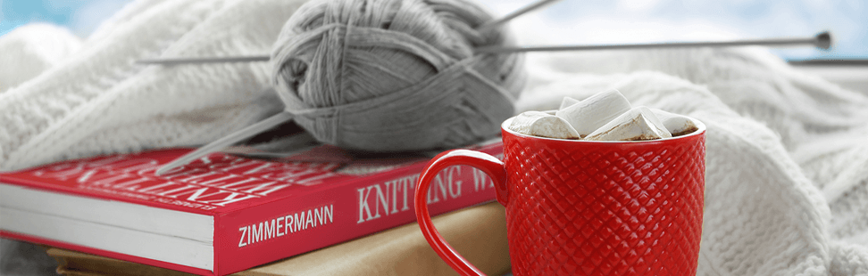 Ball of grey yarn with knitting needles on a knitting book next to a cup of hot chocolate.png