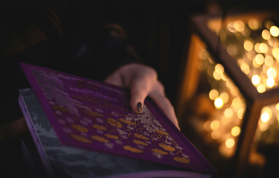 Hand holding a purple witchcraft book next to fairy lights.png