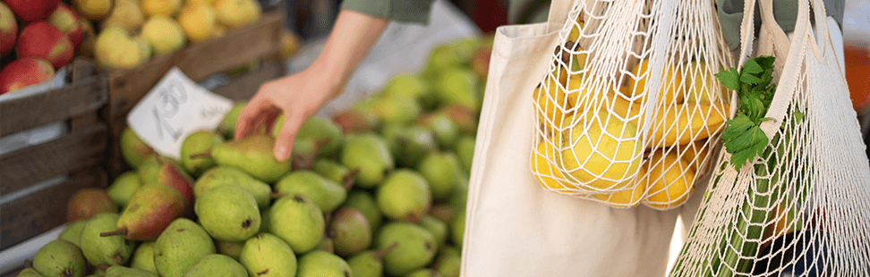 Shopper choosing pears at a market stall, holding reusable cotton and string bags of loose fruit and veg