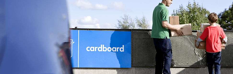A man and a boy carrying flattened cardboard boxes towards the cardboard skip at a recycling centre