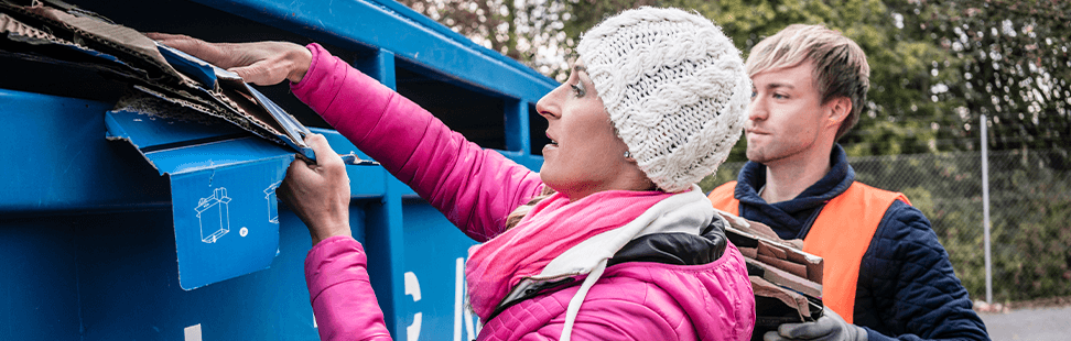 Woman posting flattened cardboard into a blue recycling skip while a man in a hi-vis vest waits with more