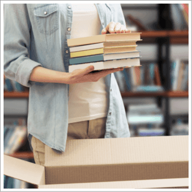 Person holding a stack of books, about to pack them into a cardboard box for selling to WeBuyBooks